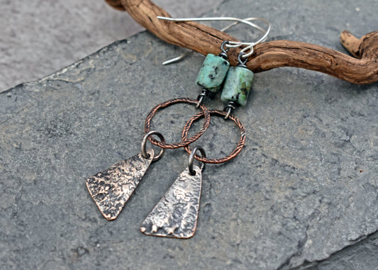 Handmade dangle earrings with turquoise beads, copper wire rings, copper triangle charms covered in melted sterling silver and sterling silver French ear wires, displayed on a piece of driftwood over a gray stone surface.