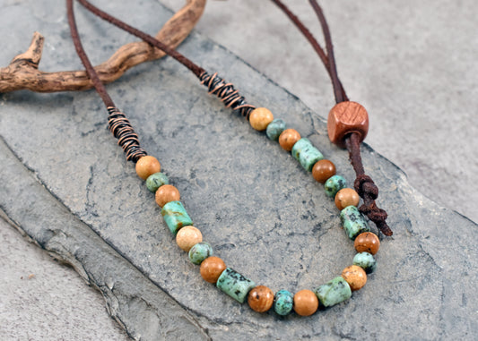 A beaded necklace with brown leather cords, featuring round and cylindrical beads in earthy brown, turquoise green, and white, displayed on a textured gray stone surface.