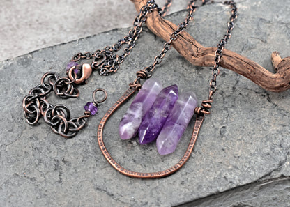 A handmade pendant necklace featuring three polished purple amethyst crystal points and copper wire resting on a gray stone surface next to a piece of driftwood.