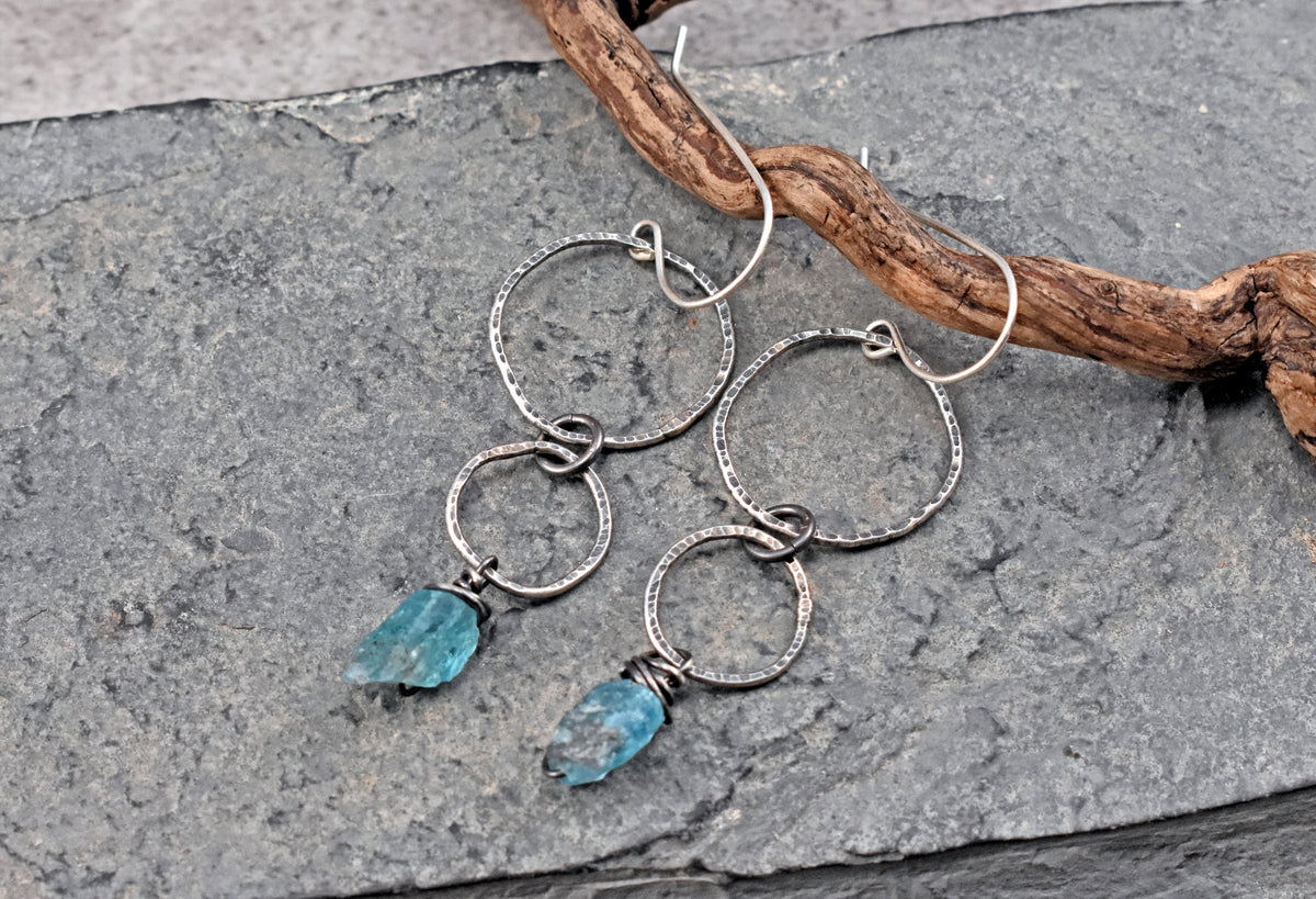 A pair of handmade dangle earrings with textured sterling silver rings and small apatite stone charms, displayed on a gray stone surface next to a piece of driftwood.