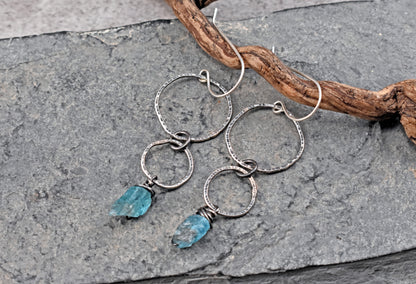 A pair of handmade dangle earrings with textured sterling silver rings and small apatite stone charms, displayed on a gray stone surface next to a piece of driftwood.