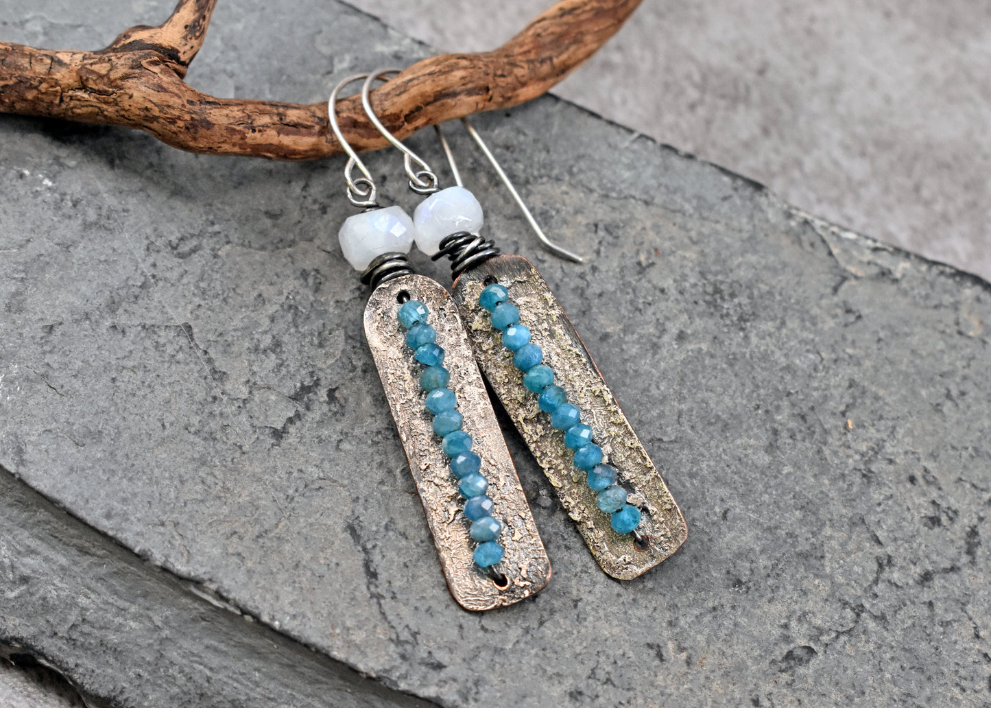 A pair of handmade dangle earrings with rectangular textured silver plates, apatite blue beads, and moonstone beads, displayed on a gray slate surface with a piece of driftwood.