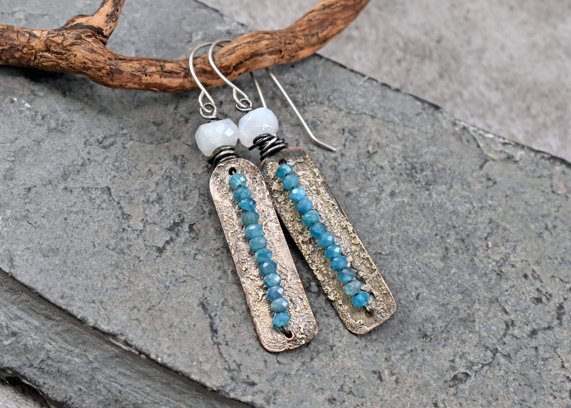 A pair of handmade dangle earrings with rectangular textured silver plates, apatite blue beads, and moonstone beads, displayed on a gray slate surface with a piece of driftwood.