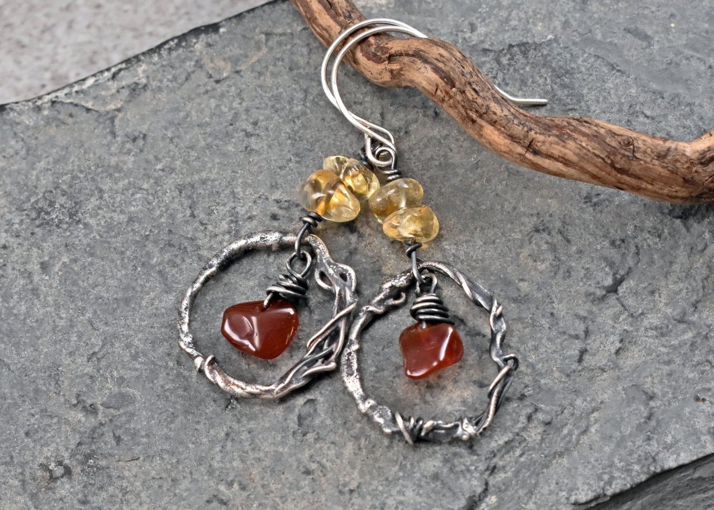 A pair of handmade dangle earrings resting on a textured gray slate surface with a piece of driftwood. The earrings feature sterling silver twisted wire hoops, small yellow citrine and red carnelian beads. 