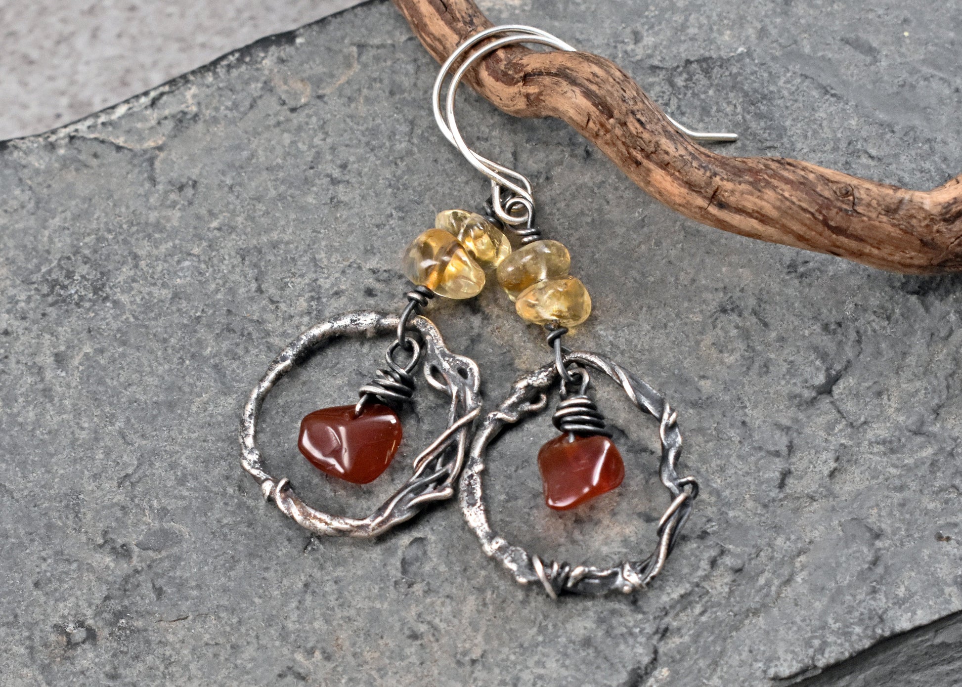 A pair of handmade dangle earrings resting on a textured gray slate surface with a piece of driftwood. The earrings feature sterling silver twisted wire hoops, small yellow citrine and red carnelian beads. 