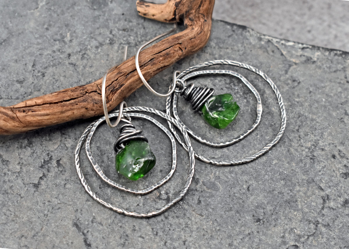 A pair of handmade dangle earrings with sterling silver wire loops and green raw chrome diopside beads, displayed on a textured gray slate surface next to a piece of driftwood.