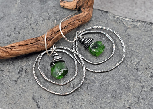 A pair of handmade dangle earrings with sterling silver wire loops and green raw chrome diopside beads, displayed on a textured gray slate surface next to a piece of driftwood.