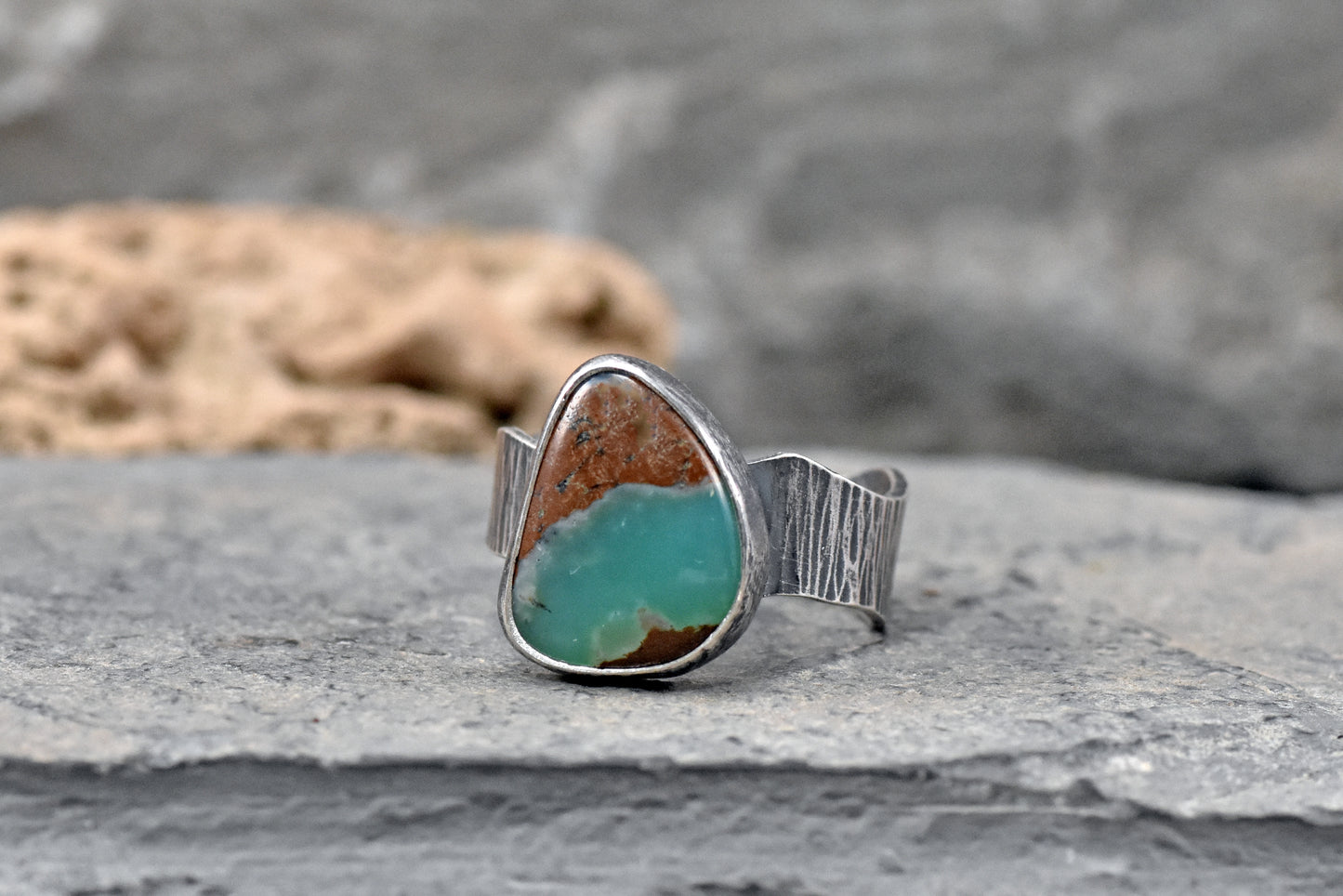 A sterling silver ring featuring a green  chrysoprase stone with natural brown matrix, resting on a gray stone surface with a blurred natural background.