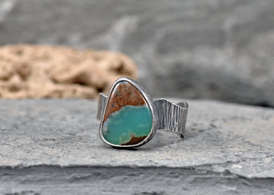 A sterling silver ring featuring a green  chrysoprase stone with natural brown matrix, resting on a gray stone surface with a blurred natural background.