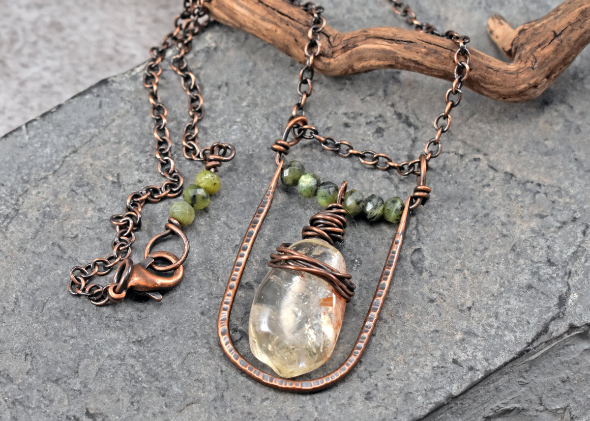 A handmade copper wire pendant necklace featuring a pale yellow citrine crystal and small jade beads, displayed on a textured gray slate surface with a piece of driftwood.