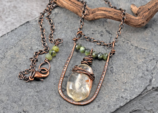 A handmade copper wire pendant necklace featuring a pale yellow citrine crystal and small jade beads, displayed on a textured gray slate surface with a piece of driftwood.