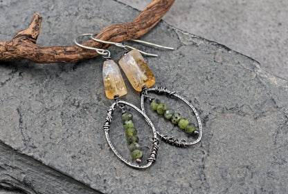 A pair of handmade dangle earrings displayed on a dark gray slate surface next to a piece of driftwood. The earrings feature yellow citrine beads and green jade bead strands wrapped in sterling silver wire.