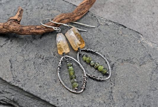 A pair of handmade dangle earrings displayed on a dark gray slate surface next to a piece of driftwood. The earrings feature yellow citrine beads and green jade bead strands wrapped in sterling silver wire.
