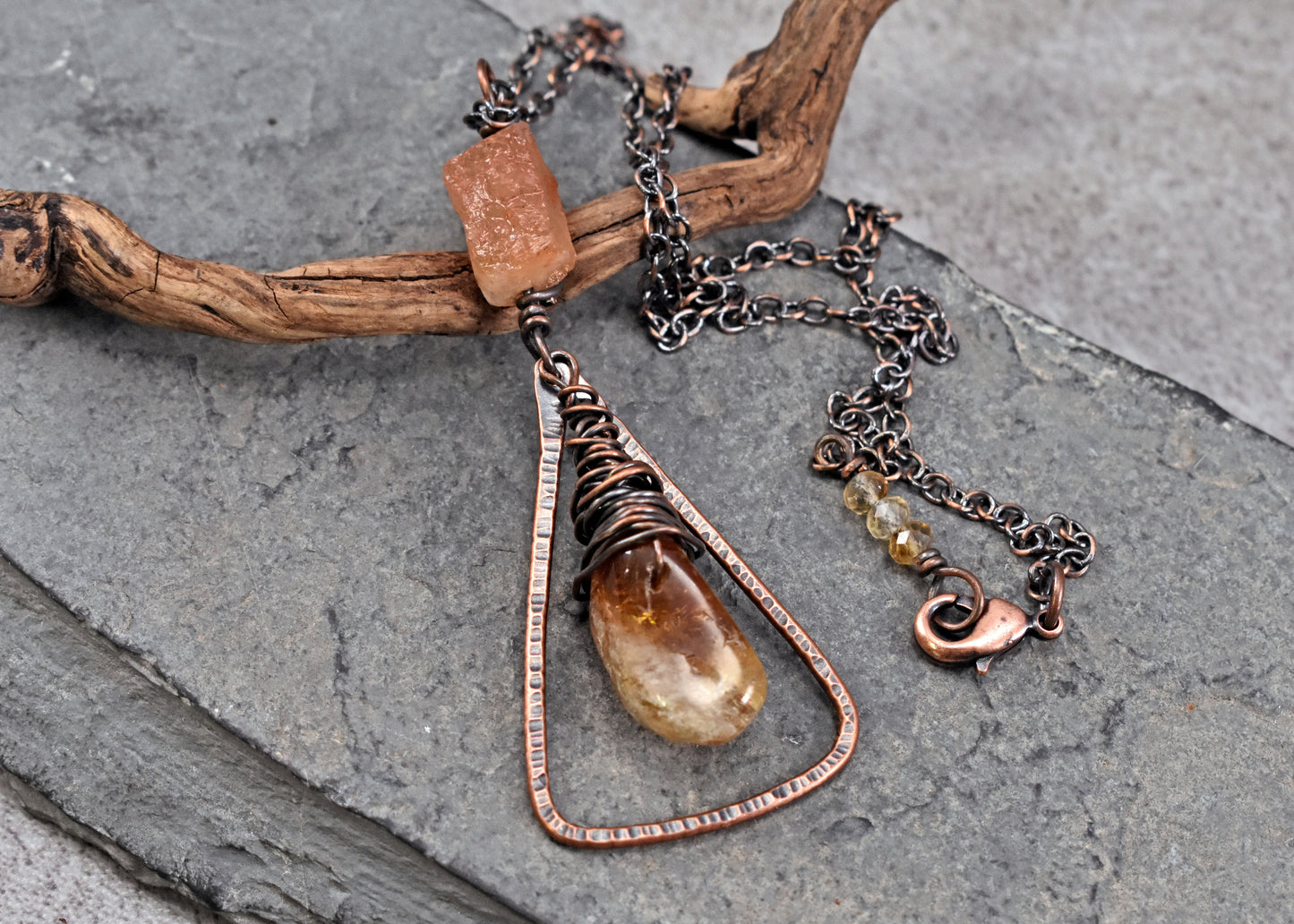 A handmade copper wire-wrapped pendant necklace with a teardrop-shaped citrine bead and orange sunstone bead, displayed on a piece of driftwood over a gray slate background.