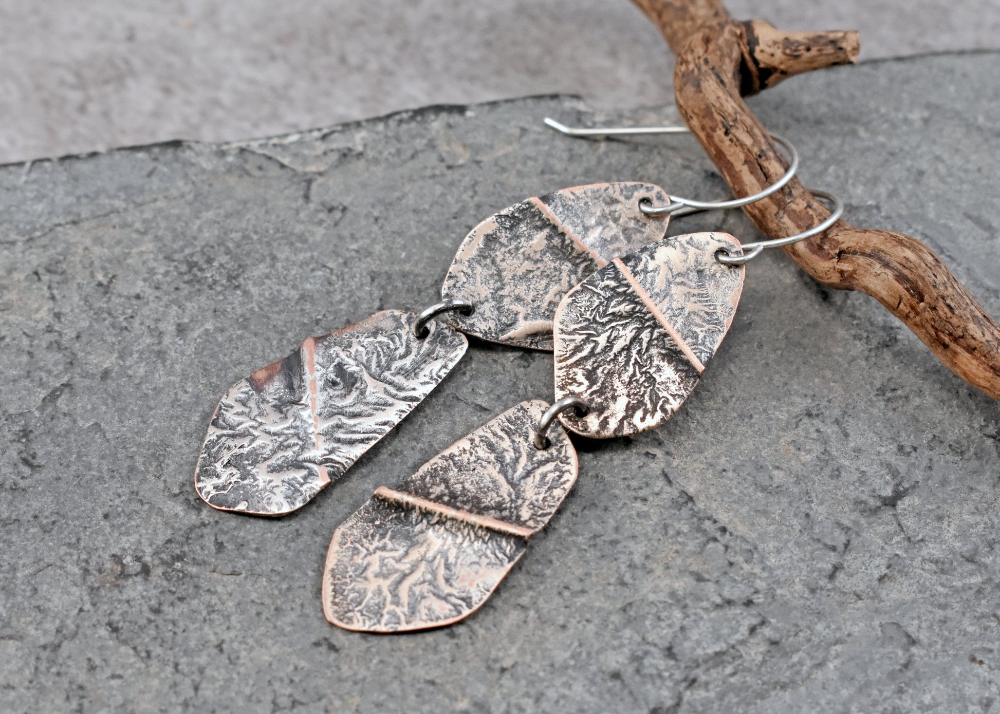 Handmade dangle earrings with textured hammered copper and sterling silver metal pieces, displayed on a gray slate surface next to a piece of driftwood.