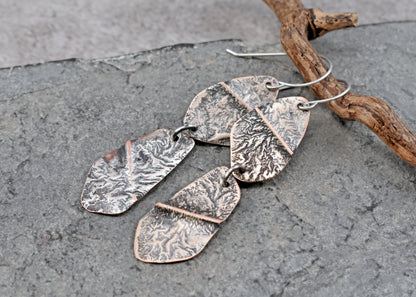 Handmade dangle earrings with textured hammered copper and sterling silver metal pieces, displayed on a gray slate surface next to a piece of driftwood.