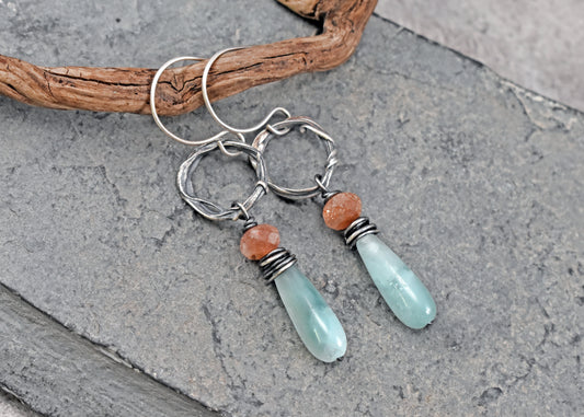 A pair of handmade dangle earrings featuring light green teardrop beads, small orange sunstone accents, and silver wire-wrapped hoops, displayed on a gray slate surface with a piece of driftwood.