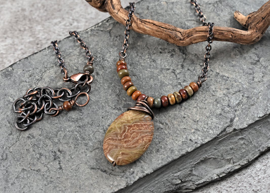 A handmade bohemian-style copper pendant necklace with a large jasper oval stone featuring a swirling wood grain pattern, displayed on a gray slate surface next to a matching chain accessory with interlocking links.