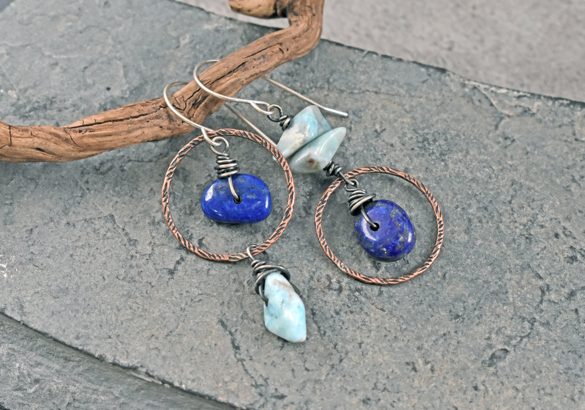 A pair of handmade dangle earrings with copper wire hoops, blue lapis lazuli stones, and light blue larimar stones, displayed on a gray slate surface with a piece of driftwood.