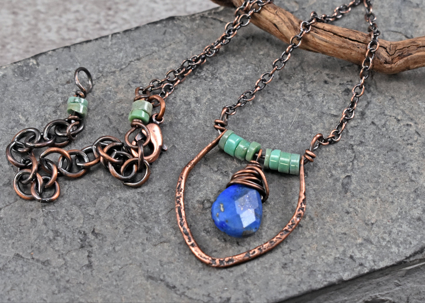 Handmade copper necklace with a blue  lapis lazuli faceted stone pendant and green chrysoprase beads, displayed on a gray slate surface with a piece of driftwood.