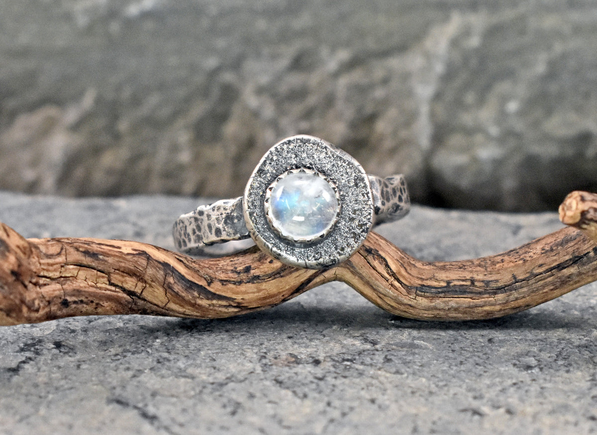 A sterling silver ring with a round iridescent moonstone center stone and a halo of texture silver displayed on a piece of driftwood against a gray stone background.