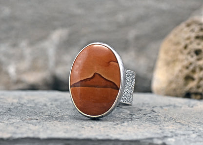 An oval sterling silver ring featuring a polished picture jasper stone with a mountain-like pattern, displayed on a textured gray stone surface.
