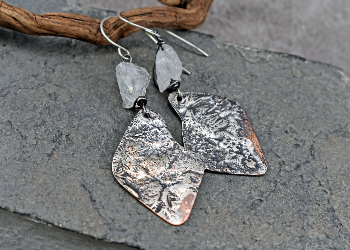 Handmade copper dangle earrings with melted silver and raw quartz stones displayed on a dark gray slate surface with a piece of driftwood.