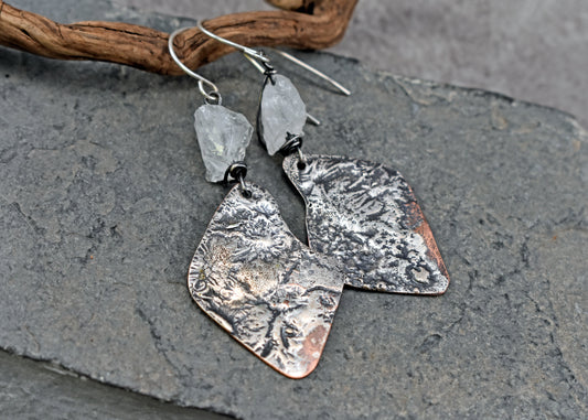 Handmade copper dangle earrings with melted silver and raw quartz stones displayed on a dark gray slate surface with a piece of driftwood.