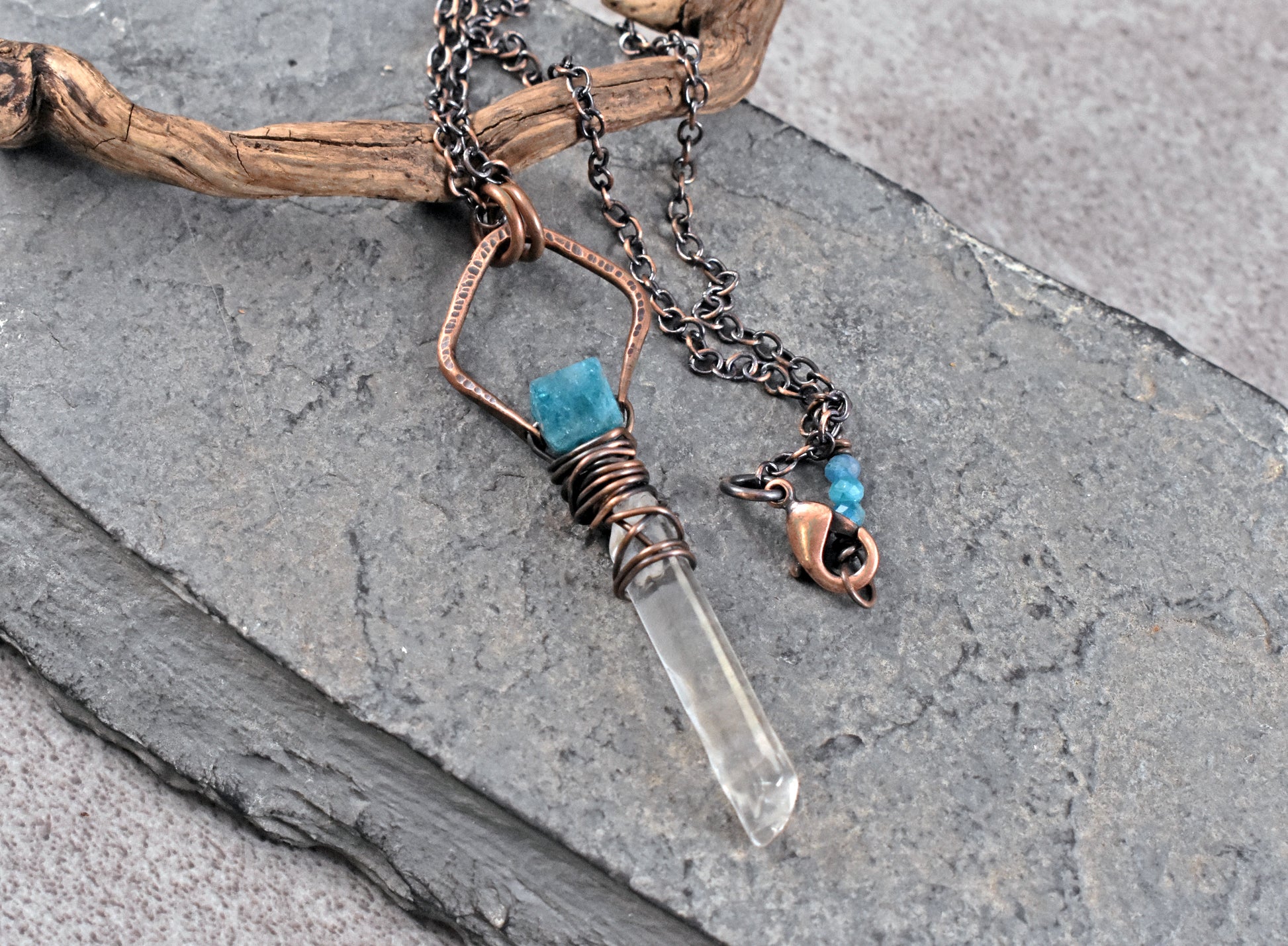A handmade copper pendant necklace featuring a clear quartz crystal point, a small apatite bead, and a copper charm, displayed on a piece of driftwood over gray slate.
