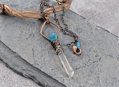 A handmade copper pendant necklace featuring a clear quartz crystal point, a small apatite bead, and a copper charm, displayed on a piece of driftwood over gray slate.