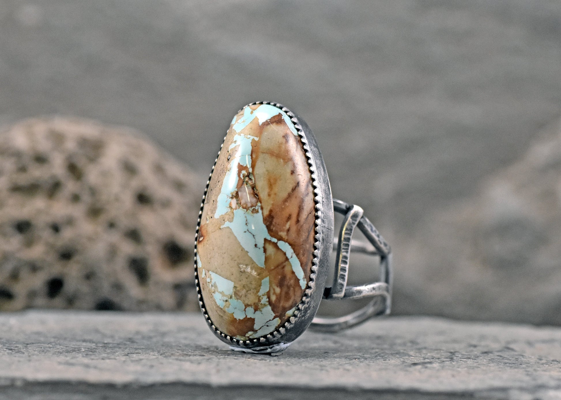 A sterling silver ring featuring a large, teardrop-shaped turquoise stone with brown and cream matrix patterns, resting on a textured gray stone surface.