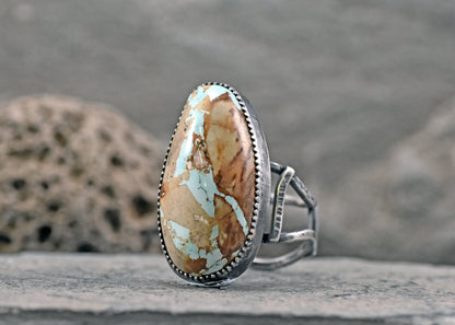 A sterling silver ring featuring a large, teardrop-shaped turquoise stone with brown and cream matrix patterns, resting on a textured gray stone surface.
