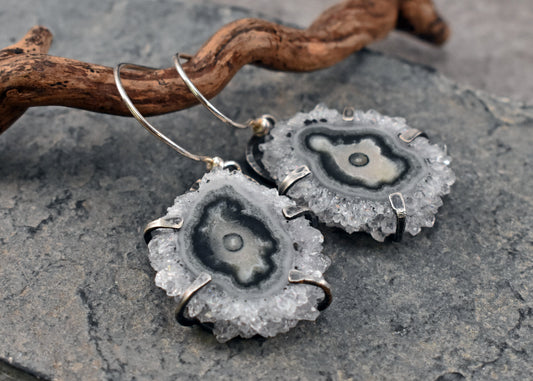 A pair of sterling silver dangle earrings featuring natural druzy agate crystal discs with black and gray banded patterns, resting on a textured gray stone surface next to a piece of driftwood.