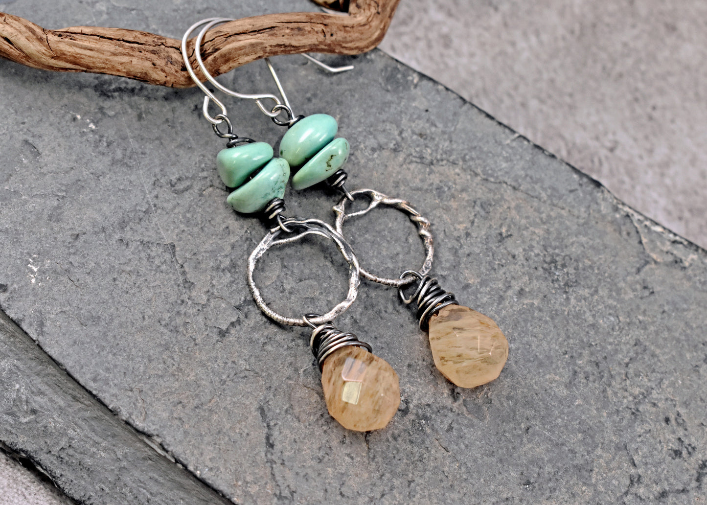 A pair of handmade dangle earrings featuring light green turquoise beads, silver wire-wrapped hoops, and faceted pale yellow quartz drops, displayed on a textured gray slate surface with a piece of driftwood.