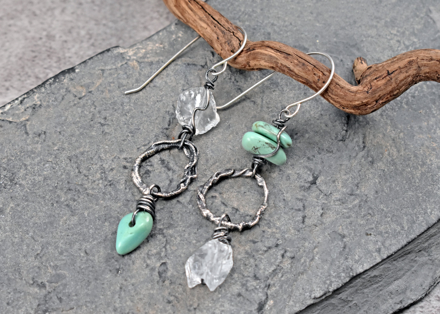 A pair of handmade dangle earrings with sterling silver rings, light green turquoise stone beads and clear quartz crystals, displayed on a piece of driftwood over a gray slate surface.