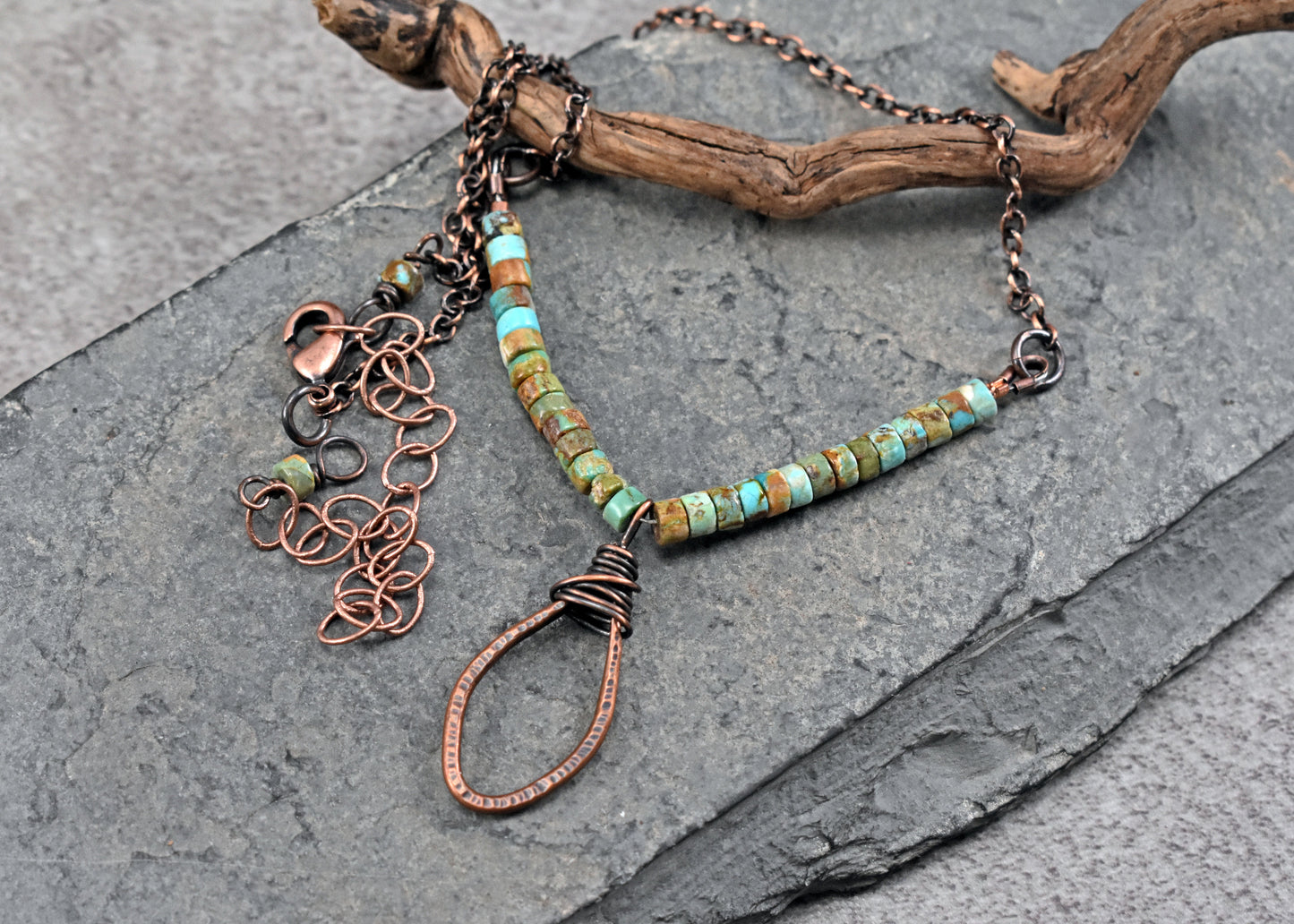 A handmade copper wire pendant necklace with turquoise beads, displayed on a dark gray slate surface with a piece of driftwood.