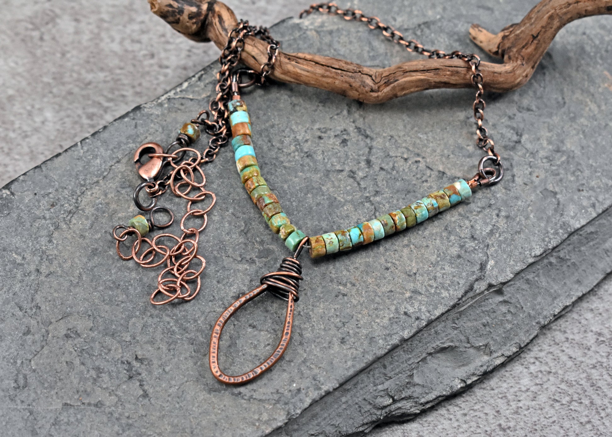 A handmade copper wire pendant necklace with turquoise beads, displayed on a dark gray slate surface with a piece of driftwood.