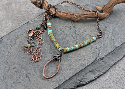 A handmade copper wire pendant necklace with turquoise beads, displayed on a dark gray slate surface with a piece of driftwood.