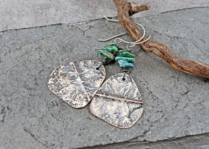 Handmade dangle earrings with textured copper and sterling silver leaf-shaped pendants, green turquoise beads, and silver fishhook wires, displayed on a gray stone surface next to a piece of driftwood.